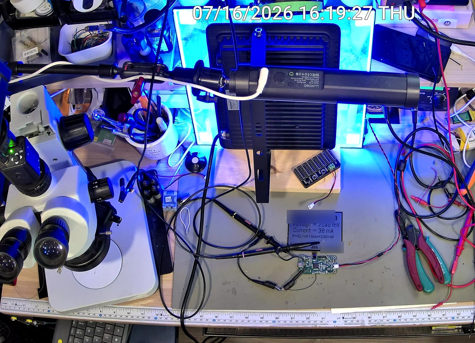 The glass panel lit under a lamp on the bench, its display reading 2140 millivolts and 38 milliamps, with oscilloscope probes on the board; the name on the display is blurred.