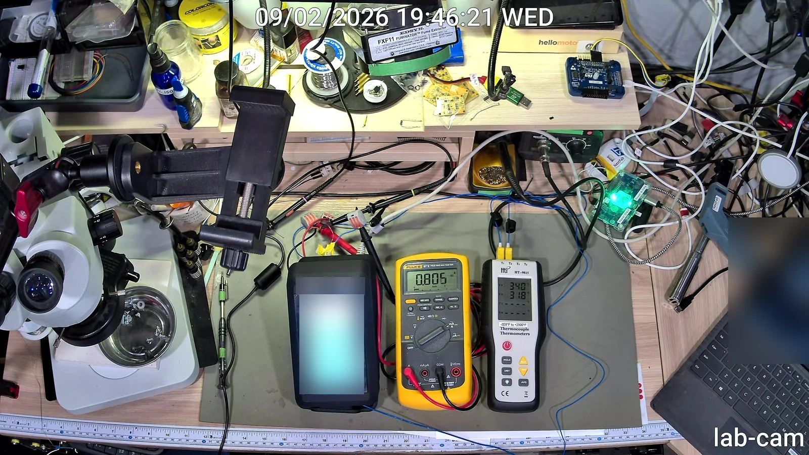 The instrument on the bench between a multimeter reading 0.805 amps and a thermocouple meter; the device screen is blurred.